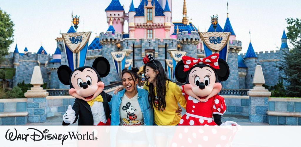 A group is happily posing in front of a colorful castle at a theme park. The two people in the middle are smiling widely, while on either side, two characters in costumes with large ears, one in red with polka dots and the other in red shorts, join the fun. The background features vibrant blue and pink turrets, creating a magical setting. Lush greenery surrounds the scene, enhancing the enchanting atmosphere.