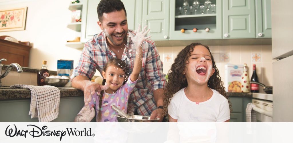 A joyful family moment in a cozy kitchen. A man with a plaid shirt is smiling while two young children enjoy baking. One child is wearing a purple outfit with stars, gleefully playing with flour. The other child, in a white shirt, laughs heartily. The kitchen has light green cabinets and various items on the countertop, creating a warm and inviting atmosphere. Walt Disney World branding is present at the bottom of the image.