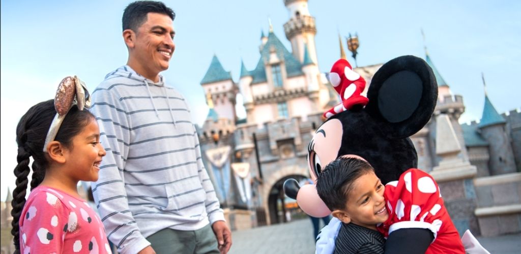 A smiling family enjoys a magical moment in front of a classic castle backdrop. A child is warmly hugged by a large cartoon character with polka-dot accessories. The joyful atmosphere captures the excitement of a theme park visit, creating lasting memories for the family.