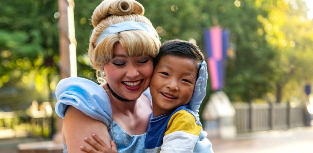 A smiling character in a light blue dress and headband embraces a smiling child outdoors. They appear joyful and are surrounded by a blurred green and purple background, suggesting a park or amusement setting. The atmosphere is bright and sunny, capturing a moment of happiness.