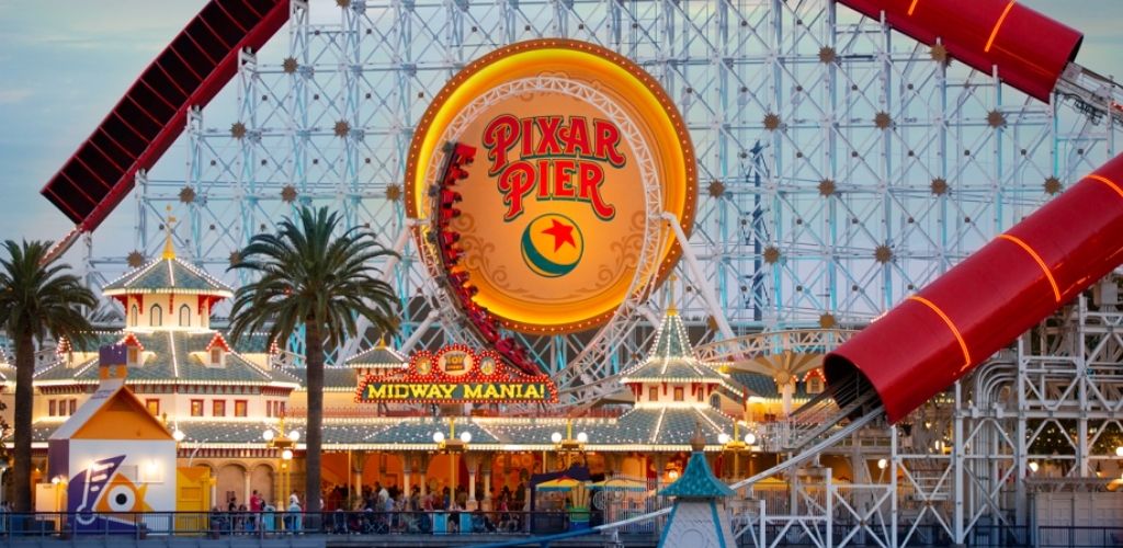 The image shows a lively amusement park area with a large ferris wheel and roller coaster. The glowing Pixar Pier sign takes center stage, surrounded by colorful lights and whimsical architecture. Palm trees line the front, and a sign for a ride called Midway Mania is visible. Visitors are seen enjoying the attractions in the vibrant, themed environment at dusk.