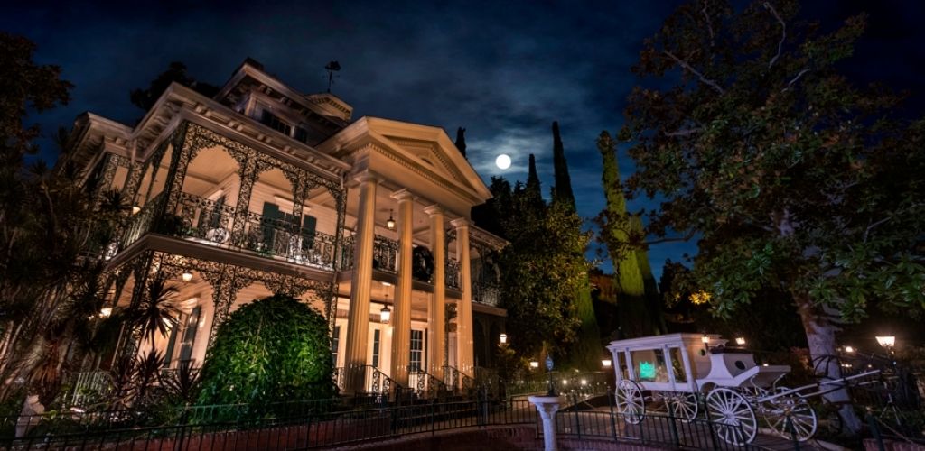Victorian-style mansion at night with glowing windows and ornate ironwork. A full moon shines through a cloudy sky, creating an eerie atmosphere. Large trees surround the house, and an old-fashioned white carriage is visible in the foreground. The scene is softly illuminated, enhancing the spooky ambiance.