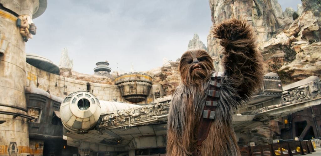 A tall, furry character resembling Chewbacca from Star Wars raises one arm in greeting. Behind him, the Millennium Falcon is parked in a detailed sci-fi setting with rugged rock formations and futuristic structures, creating an immersive atmosphere that blends natural and technological elements seamlessly.