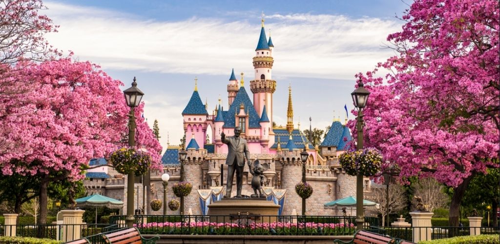 A fairytale castle with pink and blue turrets is framed by vibrant pink flowering trees. In front, a statue stands on a pedestal amid lush gardens and vintage-style street lamps. Benches offer seating areas that lead toward the whimsical structure. The sky is clear, enhancing the enchanting atmosphere.
