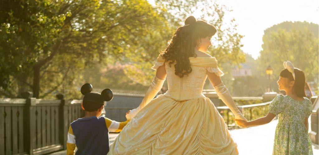 A woman in a princess costume holds hands with two children wearing mouse ears. They are walking along a sunlit path with trees in the background, creating a magical atmosphere. The image conveys a sense of joy and adventure.