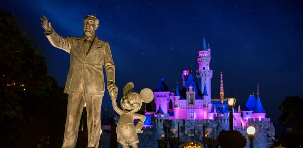 A bronze statue of a man holding hands with a mouse stands prominently in front of a beautifully illuminated castle at night. The castle glows in shades of pink and blue, creating a magical atmosphere under the starry sky. Streetlamps add a warm glow, enhancing the enchanting scene.