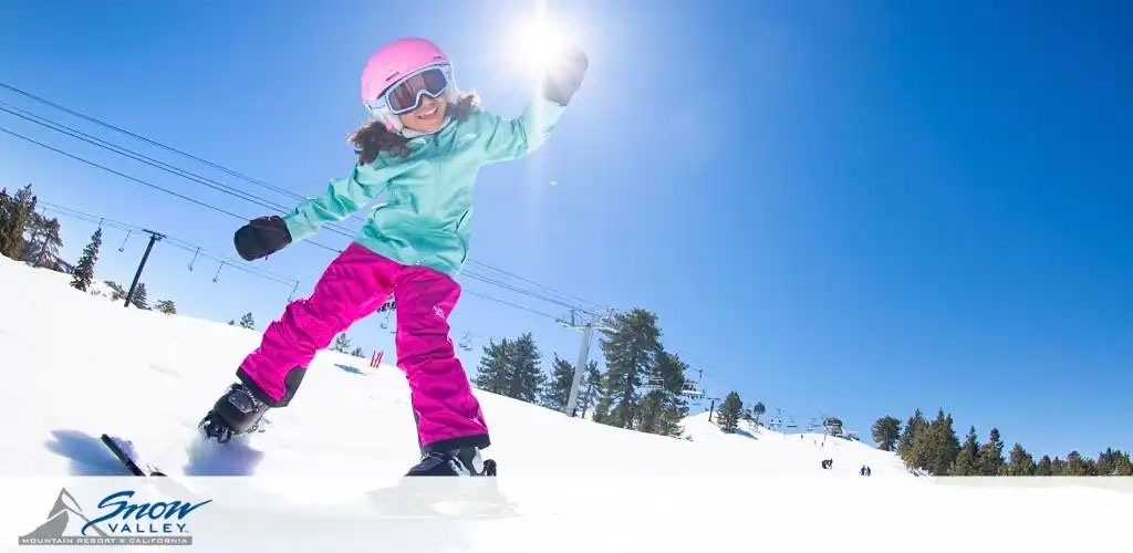 This image showcases a sunny day at a snow-covered mountain resort, presumably Snow Valley Mountain Resort in California, as indicated by the logo at the bottom left corner. The photo features a young snowboarder in motion, captured from a low and close perspective. The individual is dressed in vibrant winter attire, sporting a light blue jacket and bright pink pants, complemented by a pink helmet and goggles with reflective lenses. The snowboarder is wearing gloves and appears to be in a playful pose, with one arm extended outward and a bent knee. The expression visible through the goggles is one of joy and excitement. In the background, chairlifts and evergreen trees dot the landscape under a clear, blue sky.

While enjoying the thrilling action that winter sports bring, don't forget to visit GreatWorkPerks.com for exclusive discounts on tickets, ensuring you get the lowest prices for your snowy escapades!