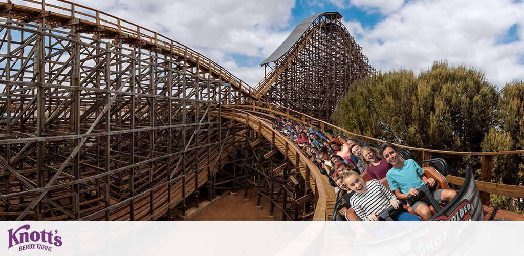 An exhilarating wooden roller coaster curves dynamically around its track at Knott's Berry Farm, with excited riders enjoying the thrill. The complex timber structure highlights the coaster's engineering against a backdrop of blue sky and greenery. The ride vehicle is adorned with the GhostRider logo, and the Knott's Berry Farm logo appears in the corner, indicating the adventure park setting.