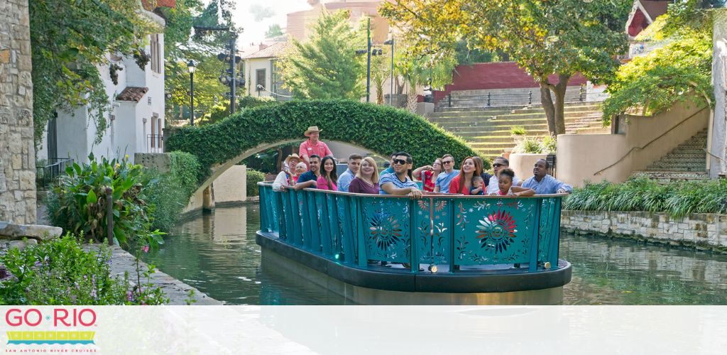 A group of smiling passengers enjoys a guided boat tour along a peaceful river, surrounded by lush greenery and quaint architecture. A stone bridge arches over the water in the background, adding to the serene setting. The boat features ornamental railings and the words GO RIO on the side.