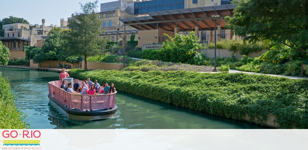 A group of people enjoys a boat tour on a calm river with lush greenery on its banks and modern buildings in the background. The boat has a pink hull with the words  GO RIO  visible on the side. Trees and a clear sky add to the pleasant atmosphere.