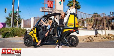 Two people are standing with a tiny yellow and black GoCar by the entrance of Boneyard Park. The person on the left is waving with a closed GoCar door behind them, and the one on the right with an open door. Both are smiling and casually dressed. The park sign and palm trees are in the background. The GoCar Tours logo is at the bottom.