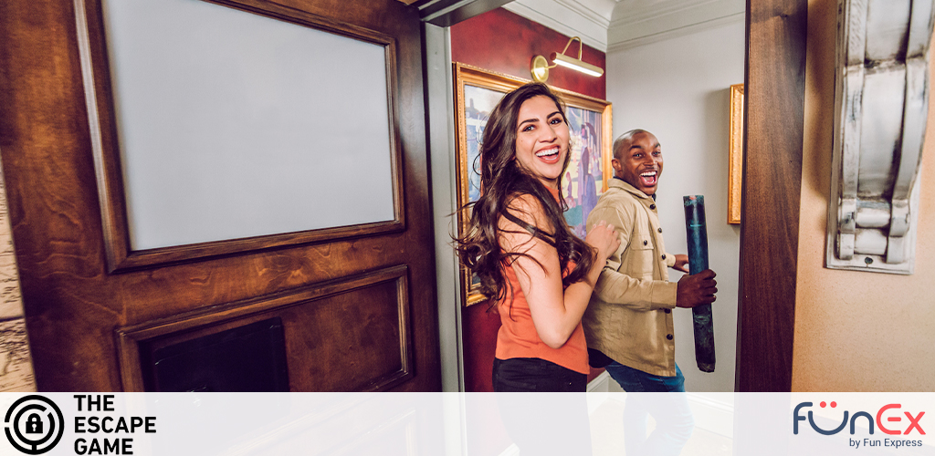 An enthusiastic woman and man appear in the middle of an escape game, working together to find clues. She is smiling broadly, looking back towards the camera, while he peers around a half-open door holding what looks to be a clue. Excitement is evident in their expressions. The setting has an old-time ambiance with wood paneling and artwork on the walls. Logos for 'The Escape Game' and 'FunEx by Fun Express' are visible in the corners.