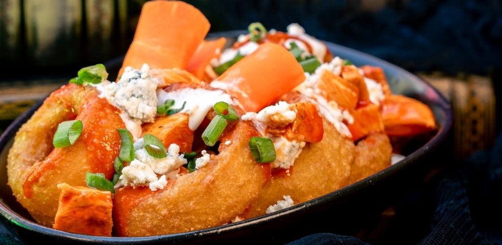 Golden fried chicken topped with spicy buffalo sauce, blue cheese crumbles, chopped green onions, and carrots on a dark plate.