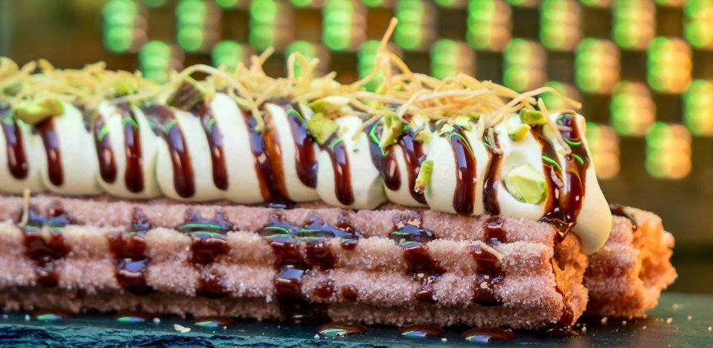 A close-up of a yummy cake with white frosting, chocolate drizzle, shredded coconut, and chopped pistachios on top.