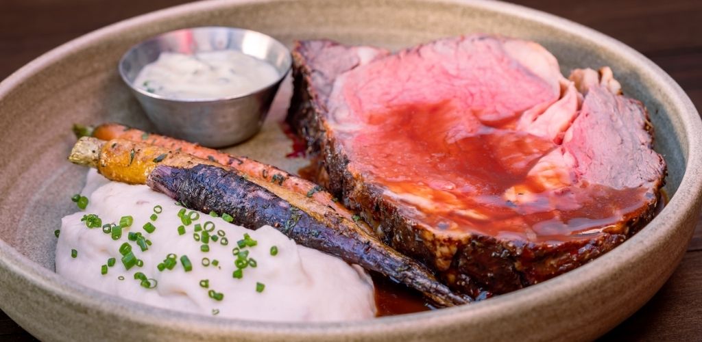 A plate with a large beef brisket, grilled salmon fillet, a serving of mashed potatoes with green onions, and a small bowl of white dipping sauce.