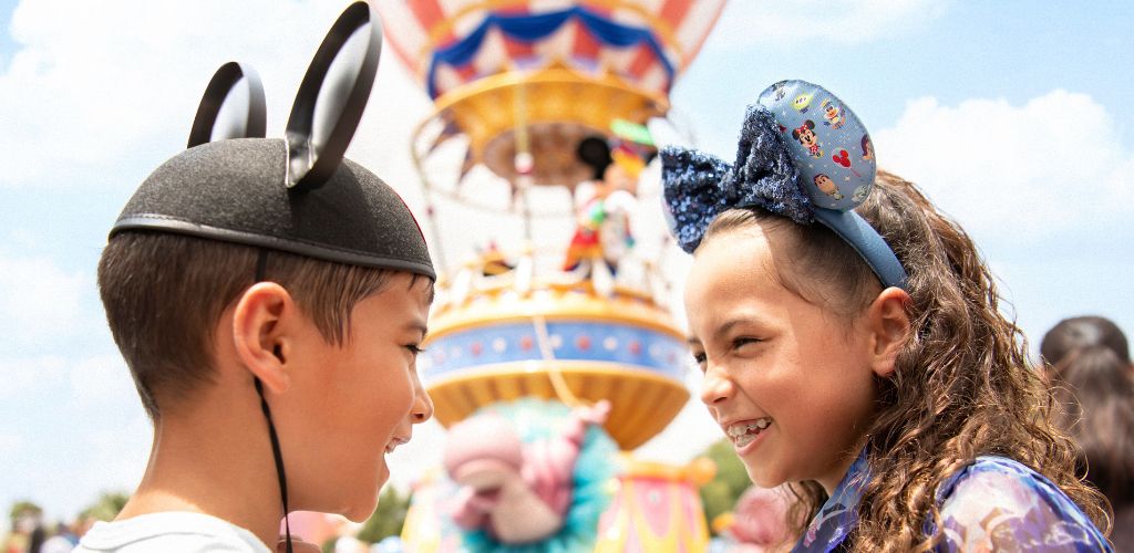 Two children face each other smiling at an amusement park with a colorful ride in the background, wearing themed headbands and enjoying a sunny day.