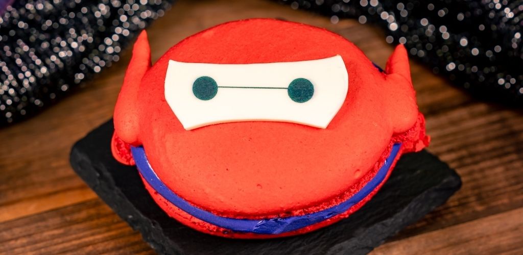 Red birthday cake shaped like a character with a white mask and black eyes, decorated with blue and red icing, on a black slate plate against a wooden background