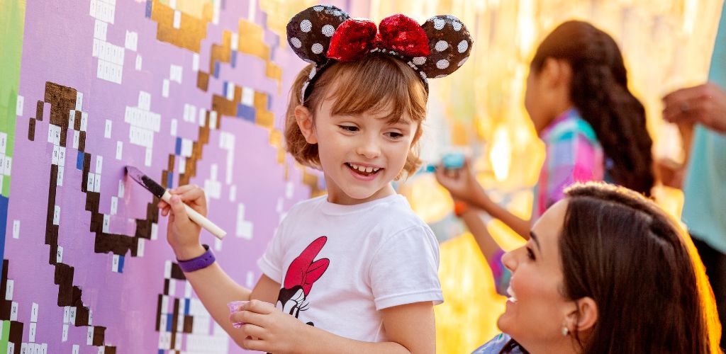 A young girl wearing Minnie Mouse ears happily creating pixel art on a colorful wall with an adult watching and smiling.