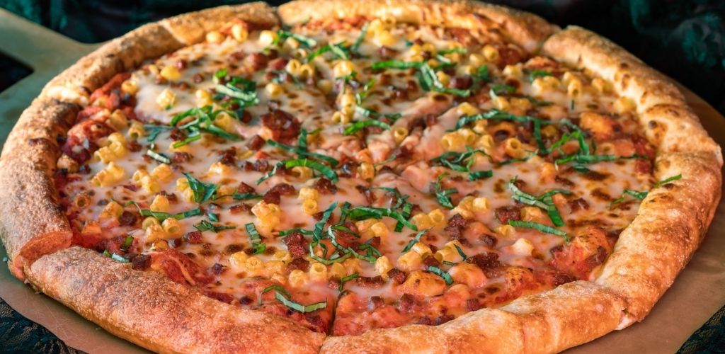 A large pizza with a thick crust topped with melted cheese, green herbs, corn, and pieces of bacon and chicken.