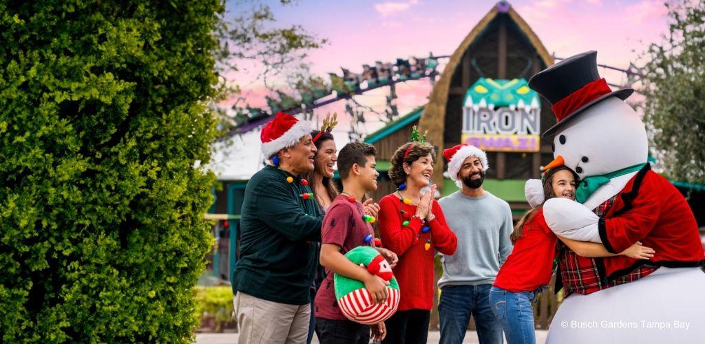 A group of joyful people in festive attire, including santa hats and reindeer antlers, share a moment with a life-size snowman figure in front of the Iron Gwazi roller coaster entrance at Busch Gardens Tampa Bay. The atmosphere is celebratory, set against a bright daytime backdrop.