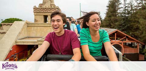 Image of a joyful young man and woman on a roller coaster, smiling and having fun. They are seated in a ride cart with other riders in the background. A blurred amusement park environment surrounds them, with a hint of greenery and structures. The logo at the bottom indicates they are at Knott's Berry Farm.