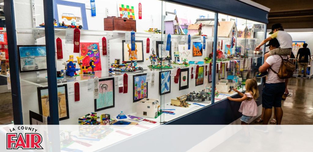 This image depicts an indoor scene at what appears to be a fair or exhibition, with a focus on a display of colorful and creative items. At the forefront, there are several enclosed display cases presenting various objects and artworks. These include LEGO models, drawings, and small constructions. Each exhibit is accompanied by descriptive tags or labels, with some items having award ribbons, suggesting a competition or judging element. A banner at the top left corner reads 'LA COUNTY FAIR'. The scene includes visitors: in the center, a man with a child on his shoulders and a young girl stand, observing the displays with interest. The overall atmosphere is engaging and family-friendly, with the visitors taking part in the interactive experience of the exhibition.

At GreatWorkPerks.com, you can find the lowest prices and significant savings on tickets for a variety of family-friendly events and fairs like the one in the image, ensuring that you don't miss out on the fun.