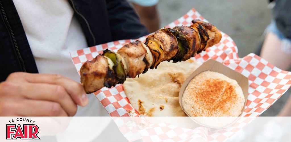 This image features a close-up view of a person holding a skewer with chunks of grilled chicken and vegetables, positioned above a paper tray with a red and white checkered pattern. Accompanying the skewer in the tray is a piece of flatbread with a sprinkle of a red seasoning. The backdrop is softly focused, highlighting the entree as the main subject. The top right corner of the image includes text that reads "LA COUNTY FAIR."

Find your next adventure and indulge in the culinary delights of the LA County Fair, all while enjoying the lowest prices on tickets when you shop with GreatWorkPerks.com.