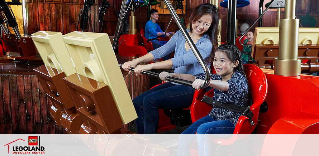 Image shows a joyful woman and girl on a red LEGO-themed ride at LEGOLAND Discovery Center. They are secured in their seats, enjoying a simulated ride with giant LEGO bricks in the background suggesting movement and fun. The setting is indoors with others visible enjoying the attractions.