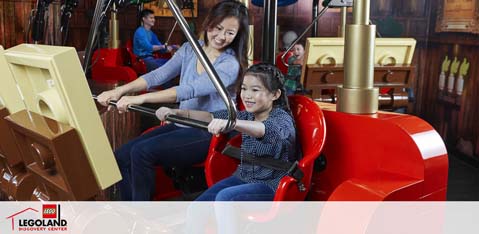 A smiling adult and child are enjoying a ride at LEGOLAND Discovery Center. They're seated in a bright red, LEGO-themed car, holding onto faux control levers, simulating an interactive experience. The background resembles a workshop with a LEGO-inspired design.
