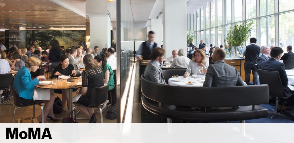 Image shows a busy restaurant scene, likely inside an art museum. Diners are seated at tables, some in conversation, while waitstaff attend to them. Natural light floods the area through large windows overlooking greenery outside. The setting conveys a modern and sophisticated atmosphere. The MoMA logo is visible at the bottom, suggesting this is the Museum of Modern Art's dining space.