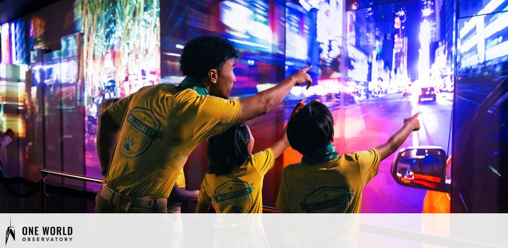 Image displaying three individuals in yellow shirts with 'VOLUNTEER' printed on the back, pointing towards neon-lit cityscape projections on a wall. The setting suggests an interactive exhibit or observatory experience, with vibrant colors and urban imagery.