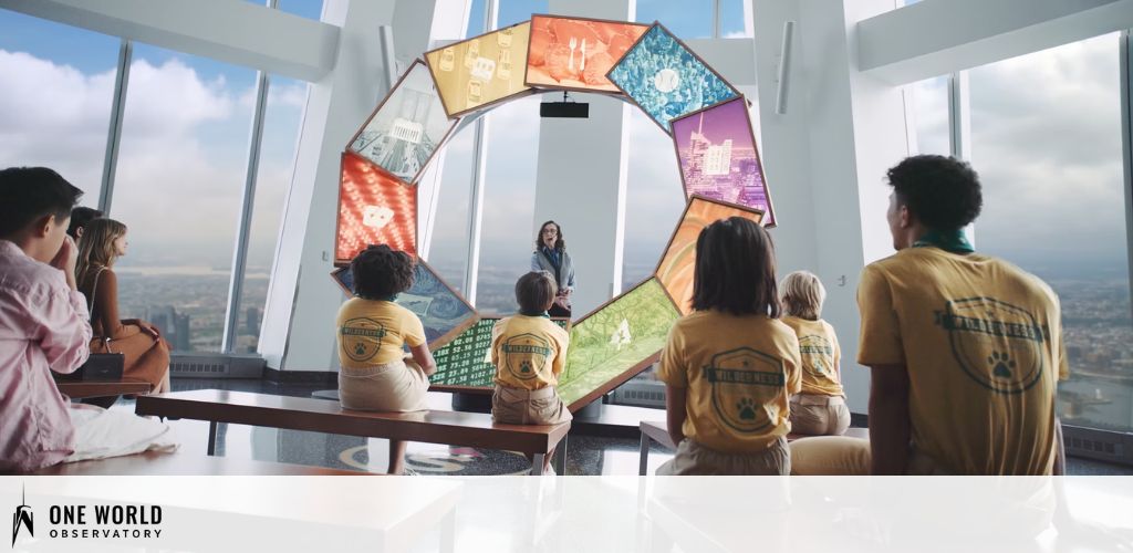 A group of visitors is seated indoors facing a presenter who stands near a display shaped like a ring with various screens. The room has large windows offering a view of the cityscape far below. The attendees appear engaged, with some wearing shirts that read One World Observatory.