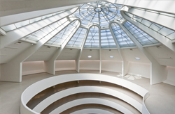 Image shows the Guggenheim Museum in New York on a sunny day. Its distinctive, curved white facade stands out against the city backdrop. People are crossing the street in the foreground, and trees with white blooms add a touch of nature to the urban environment.