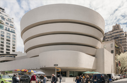 Image shows the interior of a modern building with a spiraling design. Broad, white balconies ascend around a central void towards a glass dome ceiling with a circular frame, allowing daylight to permeate the space.