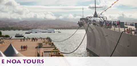 Image of a large battleship docked at a pier with visitors walking on its deck. In the distance, hills and a cloudy sky can be seen. The foreground shows a banner reading 'E NOA TOURS' on the bottom right corner.