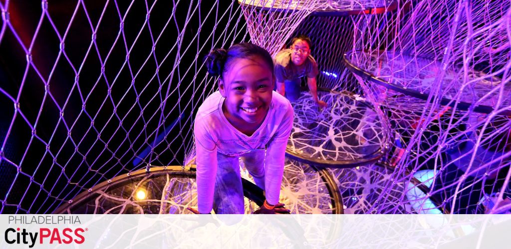 Image shows two children playing in an indoor mesh play structure with vibrant purple lighting. There's joy on the face of the child in the foreground. The Philadelphia CityPASS logo is visible in the corner, indicating an attraction included in the pass.