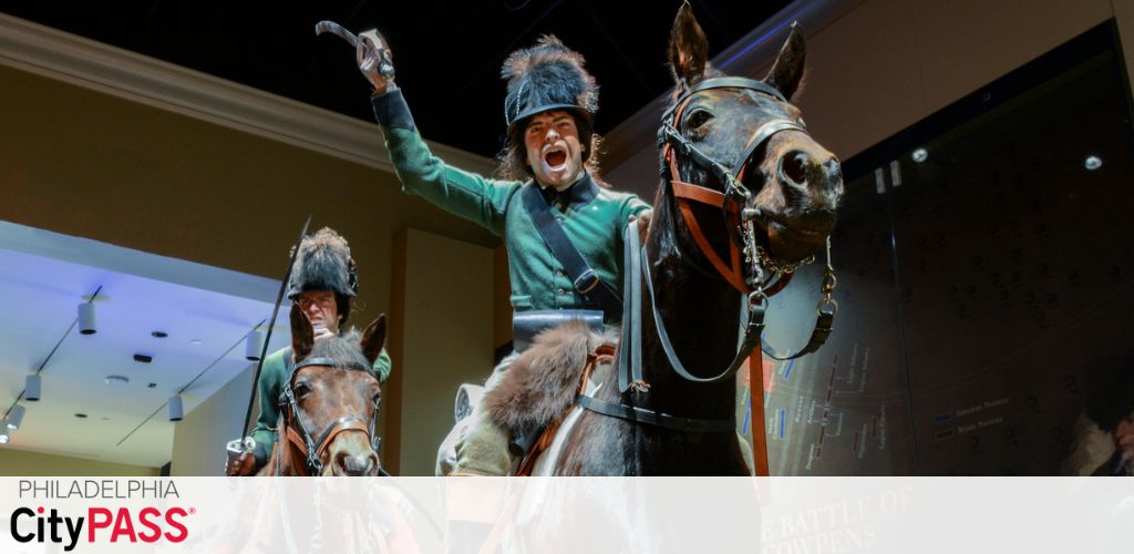 Promotional banner for Philadelphia CityPASS with a dynamic display of historical reenactment figures. The photo captures two lifelike statues of soldiers on horseback in mid-battle. The foremost figure is raising his arm in a battle cry.