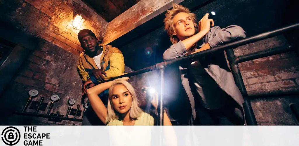 Image Description: This is a dynamic promotional photo representing The Escape Game experience. It features three individuals engaged in what appears to be an escape room scenario. On the left, a person in a yellow jacket looks on thoughtfully with their hand resting on their chin. The middle person, with blonde hair, is crouched while looking upward with an intense gaze, with one hand holding their head. On the right, another individual is gripping a railing and peering pensively into the distance, holding a flashlight that illuminates the scene. They are all surrounded by dim lighting which adds to the ambiance of mystery and discovery, with the brickwork of the room visible in the background, suggesting an underground or old-fashioned setting. The logo of The Escape Game is visible on the bottom-left corner of the image.

Integrate GreatWorkPerks.com into your escape plans for an adventurous outing, and uncover substantial savings with our lowest prices on tickets!