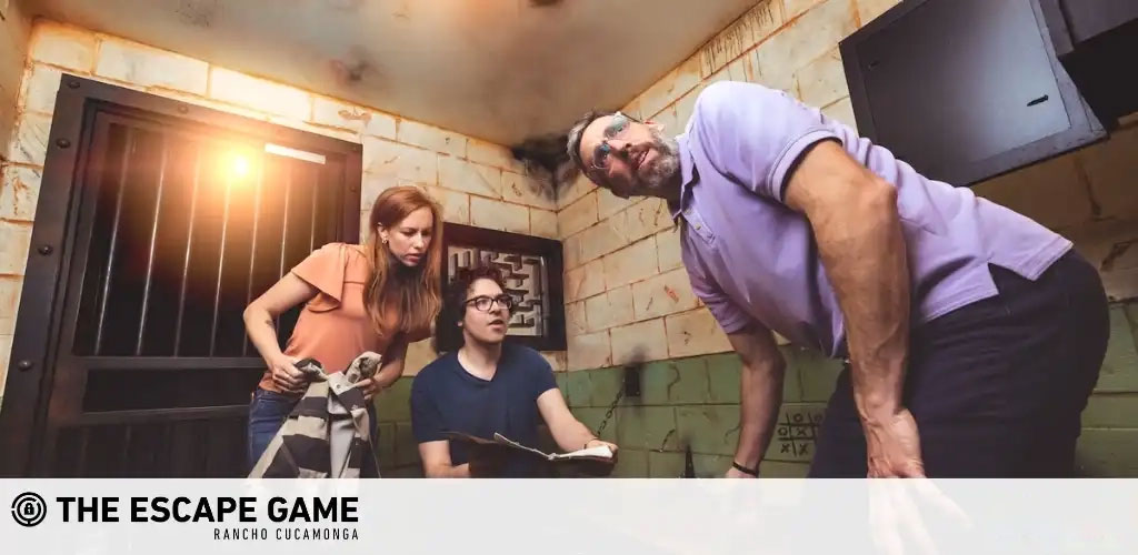 Image Description: Three individuals appear deeply engaged in an escape room challenge at The Escape Game in Rancho Cucamonga. The room features a gritty, weathered wall, and a barred window emitting a warm glow. On the left, a woman with long auburn hair, wearing an orange top, concentrates while holding an object that might be a clue. She is collaborating with a man in the center, who is wearing glasses and a blue shirt and is seated, looking upwards, possibly communicating with the standing man on the right. The man on the right has short, dark hair and is wearing a light purple shirt. He is bent slightly forward, hands braced on his knees, with an expression of thoughtful determination.

Visit GreatWorkPerks.com to unlock exclusive savings on tickets to the best escape room experiences. Our commitment ensures you get the lowest prices, so you can enjoy the thrill of the escape for less!