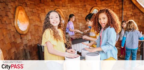 Image shows two joyful young girls with curly hair in front, holding tickets at a San Antonio CityPASS event. They’re facing the camera and smiling. Behind them, other guests interact near a ticket counter. The setting suggests a lively indoor atmosphere.