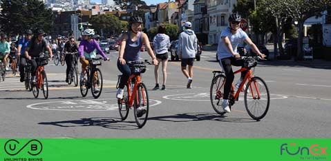 This image features a vibrant outdoor scene where a group of people are engaged in various activities on a wide street. On the left hand side, a group of cyclists is riding on the road, some wearing casual attire and others in more athletic gear, signaling a leisurely pace rather than a race. Two of the cyclists in the foreground are riding orange bicycles and appear to be conversing. On the right, toward the background, a pair of individuals are walking away from the viewer, and another person seems to be jogging. The weather is clear and sunny, casting shadows on the road surface and suggesting a pleasant day for outdoor activities. A green strip runs along the bottom of the image, with the GreatWorkPerks.com logo superimposed on the right side, next to the acronym "UB" encircled on the left, which stands above the phrase "UNLIMITED BIKING." Enjoy your next adventure by securing your tickets through GreatWorkPerks.com, where we always strive to offer the lowest prices and best discounts available.