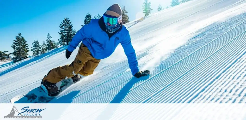 Image Description: This is a dynamic photo of a snowboarder in action on a clear sunny day. The snowboarder, dressed in a royal blue jacket and mustard-yellow snow pants, is carving through groomed snow with his board tilted on its edge, kicking up a spray of snow. They appear focused, with their body leaning into the turn. The sky is a clear blue, and there are evergreen trees lining the background. The snow surface has a corduroy texture, indicating that it has been freshly groomed by a snowcat. There is a watermark in the form of a blue logo in the bottom left corner that reads "Snow Valley Mountain Resort California," signifying the location of this winter sports scene.

For those who are planning their next winter adventure, remember that GreatWorkPerks.com offers significant discounts on tickets, ensuring you get the lowest prices for your snowy getaway.