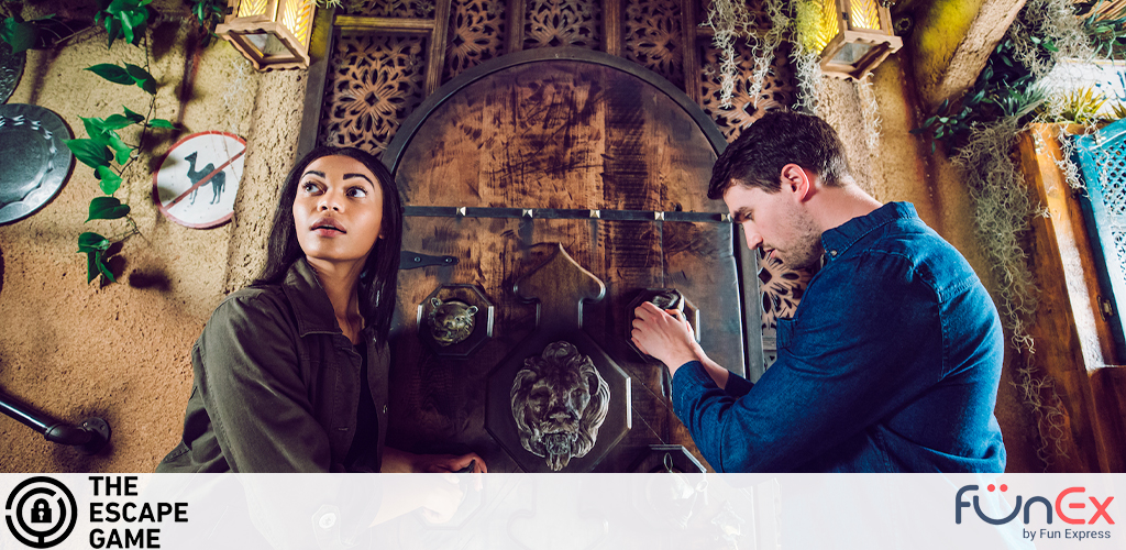 Image shows two individuals in an escape room setting, engrossed in solving puzzles. A woman looks upwards, perhaps clue searching, near a wall adorned with ivy and a no-smoking sign. A man concentrates on unlocking a box. The ambiance is adventurous with rustic decor elements. Logo watermarks present.