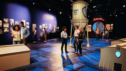 Visitors explore an exhibit at Warner Bros. Studio featuring displays, interactive kiosks, and movie memorabilia. The space is dimly lit with focus lighting, creating an immersive atmosphere.