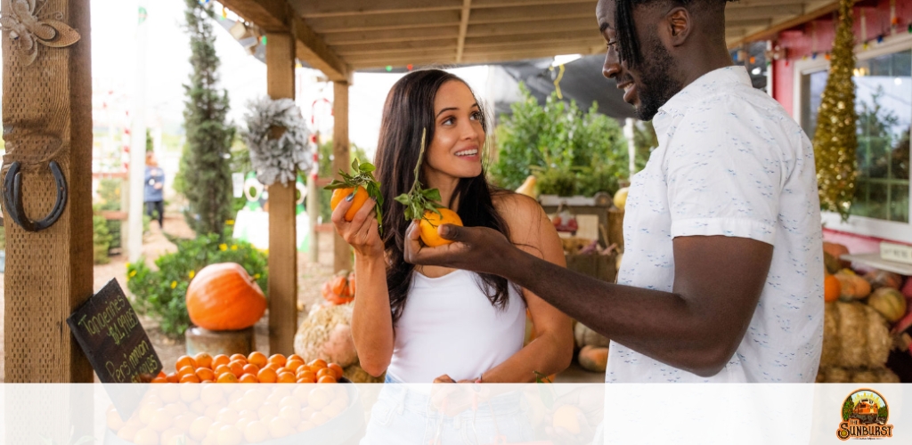 Image description: 

The photo captures a moment between two individuals, a man and a woman, at a farmers market stall. The setting appears to be outdoors under a wooden shelter, and it's daytime. The woman, to the left, holds two persimmons, one in each hand, and is looking at the man with a smile. She wears a white sleeveless top. The man, facing her and slightly turned to the right, is smiling and seems to be touching one of the persimmons she is holding, indicating a shared moment of interest in the fruit. He is wearing a white shirt with a blue feather pattern. Behind them, a table is laden with neatly arranged persimmons and there's a large orange pumpkin in the background. On the extreme left, a chalkboard sign is partially visible, with the word "Tomatoes" on it. Decorative elements like a wreath and more pumpkins suggest an autumnal theme for the market. In the background, to the left, there is another customer and some greenery, providing a pleasant ambiance. The Sunburst logo is visible in the lower-right corner of the photo.

At GreatWorkPerks.com, find the best experiences at the lowest prices guaranteed. Don't miss our exclusive discounts on tickets to a variety of fun-filled destinations!