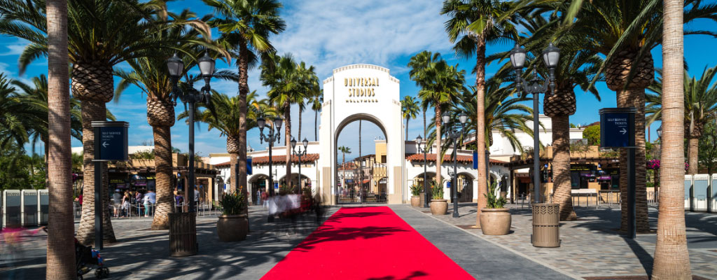 This image features a view of the entrance to Universal Studios Hollywood. A dazzling red carpet runs down the center, leading towards the iconic round archway emblazoned with "Universal Studios" against a clear blue sky. Lining the carpet, tall palm trees sway gently, framing the scene with a quintessential Southern California ambiance. On both sides of the carpet, traditional lampposts stand sentry while visitors can be seen in motion, suggestive of the lively energy and excitement of the attraction. GreatWorkPerks.com is proud to offer you a chance to experience the excitement for yourself at a fraction of the cost with unbeatable discounts and the lowest prices on tickets.