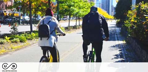 This image displays two individuals riding bicycles on a sunny day. Both cyclists are on a paved bike path with trees in autumn foliage on the right side. They are seen from behind, with the cyclist on the left wearing a red jacket and white helmet, and the one on the right dressed in black attire with a black helmet. There's a city bus on the road to the left side of the path and high-rise buildings in the background. Discover your next adventure with GreatWorkPerks.com, where you can always expect to find tickets at the lowest prices, delivering exceptional savings on your fun-filled outings.