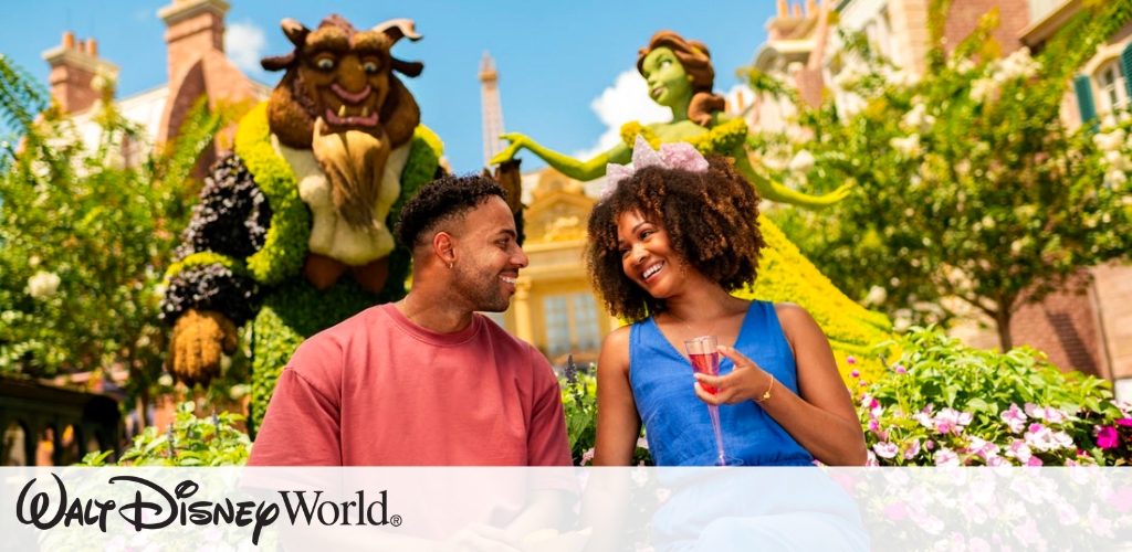 A couple sits on a bench surrounded by lush flowers and topiaries resembling fairy tale characters. The man, wearing a red shirt, and the woman, in a blue dress with a bow headband, smile at each other. Behind them are large plant sculptures of a beast and a princess. The background features a whimsical building facade. The vibrant scene exudes joy and enchantment, capturing a magical moment.