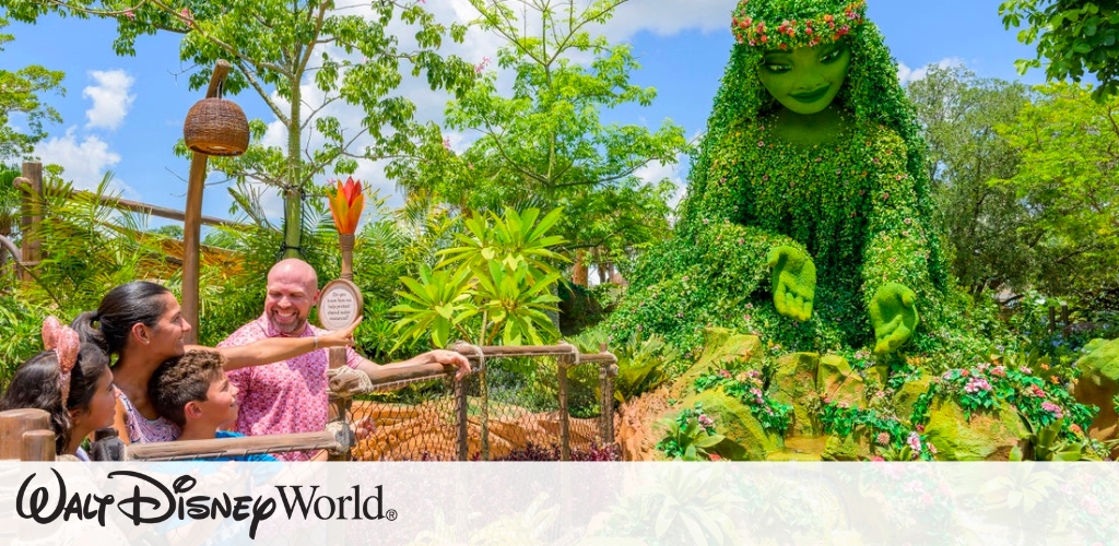 A family enjoys a sunny day at a theme park, looking at a large plant sculpture of a smiling figure adorned with a flower crown. The scene is surrounded by lush greenery and vibrant plants under a bright blue sky.