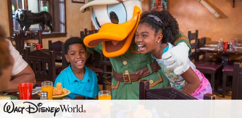 A cartoon duck character dressed as an explorer poses happily with two children, a boy and a girl, in a restaurant setting. Both children are smiling widely, sitting at a table with colorful drinks and food. The warm, inviting atmosphere suggests a fun and engaging dining experience.